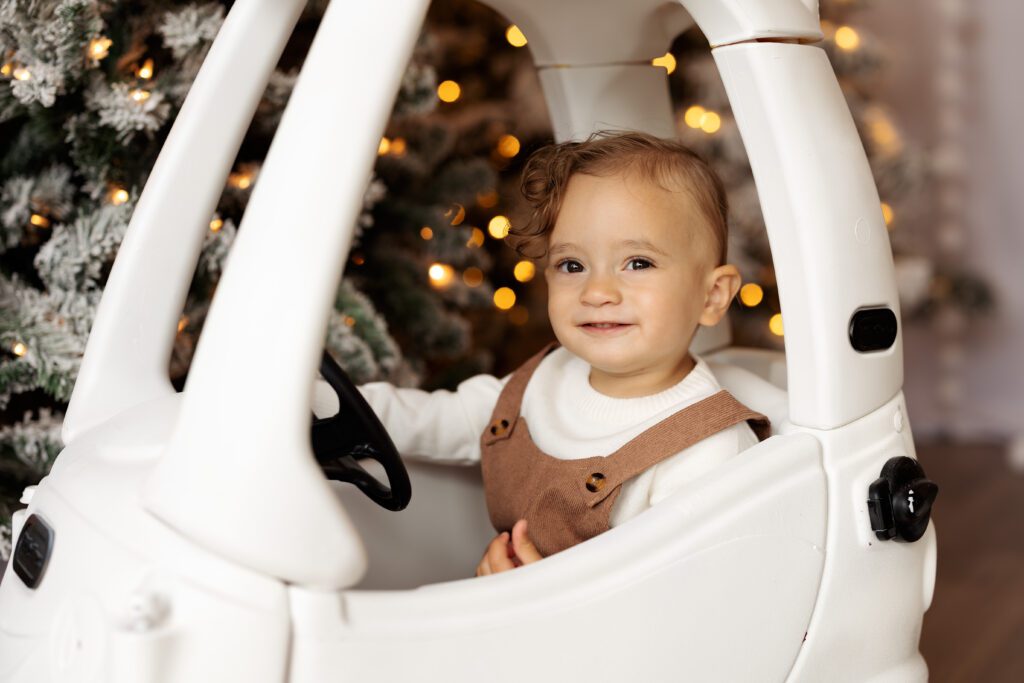 Little boy in a white little tikes car, decorated to fit in with a holiday Christmas backdrop of frosted trees. Christmas mini sessions at Kindle Studio in Colorado Springs.